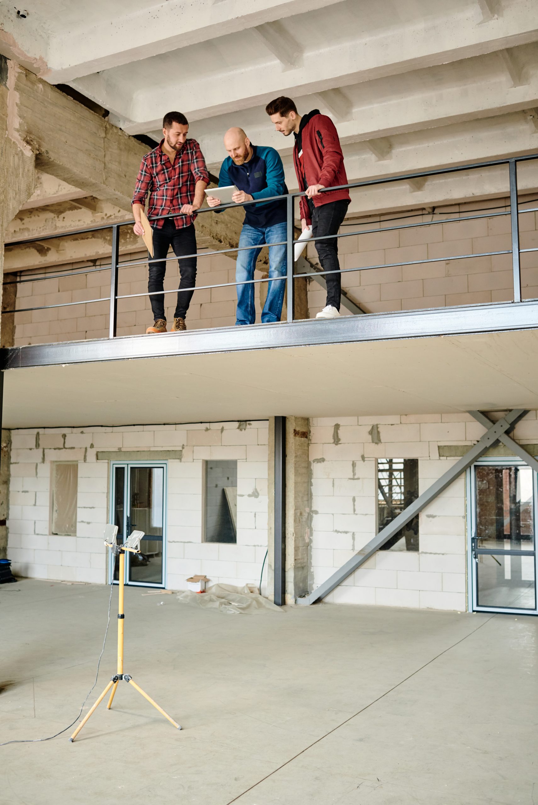 Group of three young professional architects standing by railing on the second floor of unfinished building and discussing new project